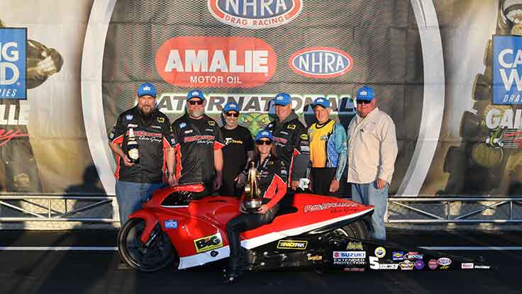 Pro Stock Motorcycle racer Karen Stoffer, sitting on motorcycle holding Wally Trophy with team.